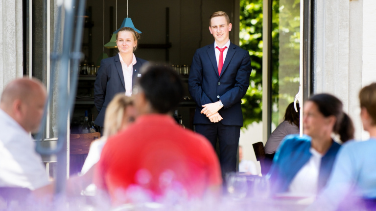 Een vrouwelijke en mannelijke eerstejaars student houden in de gaten dat het de gasten op het terras van het Teaching Hotel aan niets ontbreekt.