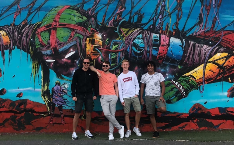 Four guys standing in front of a wall full of graffiti. Smiling and leaning on each other looking into the camera.