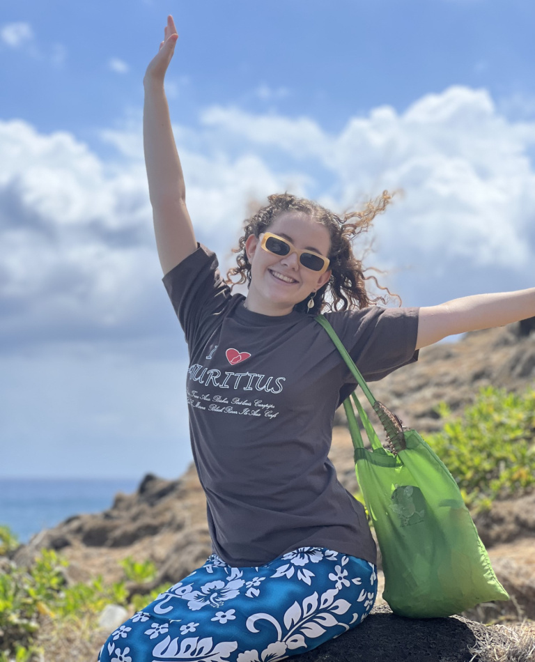 girl with curls and yellow sunglasses, wearing a brown shirt 'i love mauritius', sitting on a rock near the sea