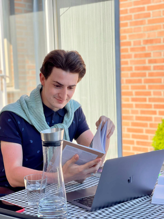 Student zit aan een tafel achter een laptop, heeft een boek in zijn handen en een karaf water op de tafel staan. Draagt een blauwe polo en een groene trui. Het tafelkleed is geblokt.