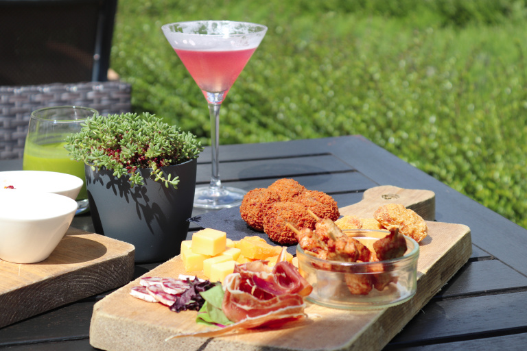 Een borrelplank op een tafel op het terras van het Teaching Hotel met een rode cocktail, bitterballen, kaas en vleeswaren,